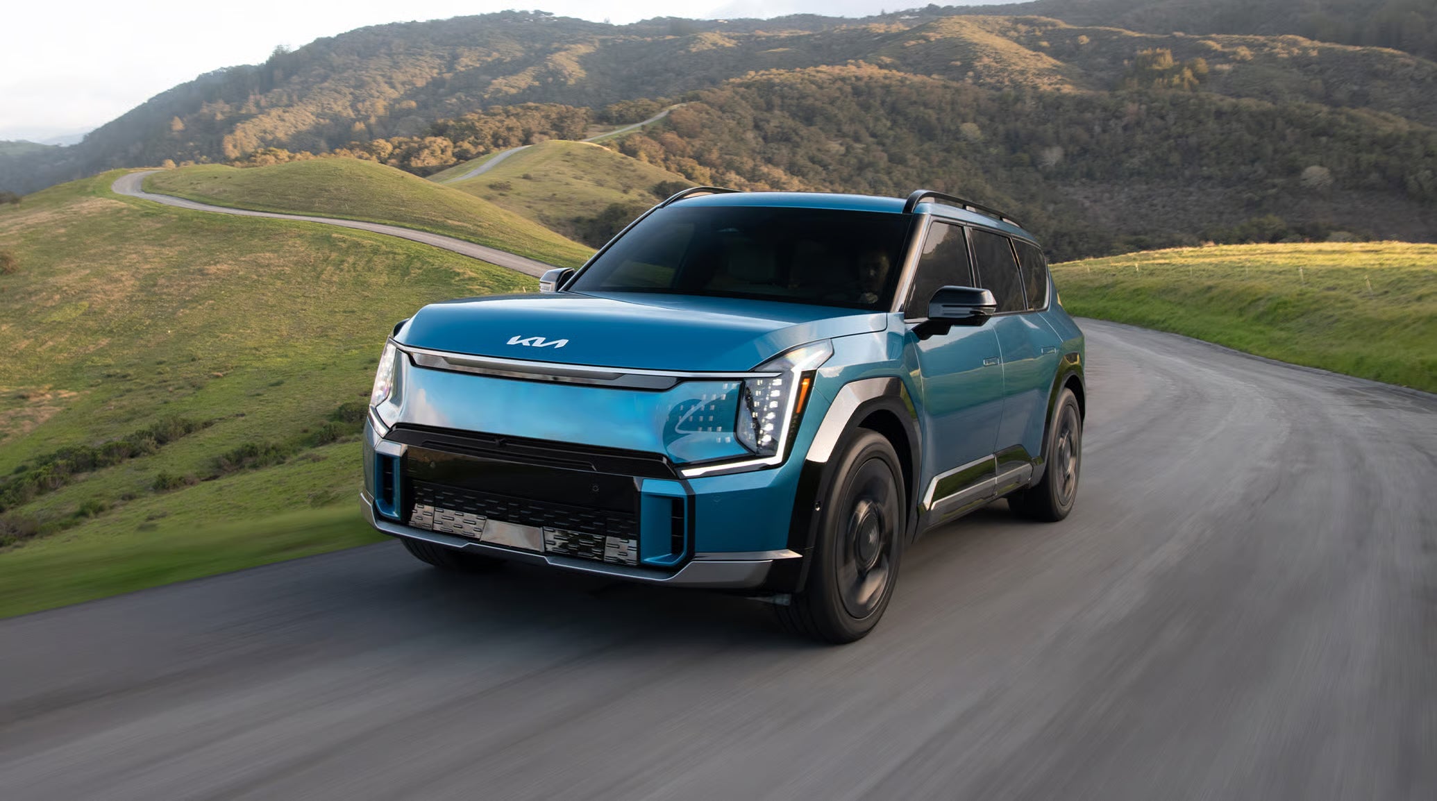 A bright blue 2026 Kia EV9 SUV driving up a hill