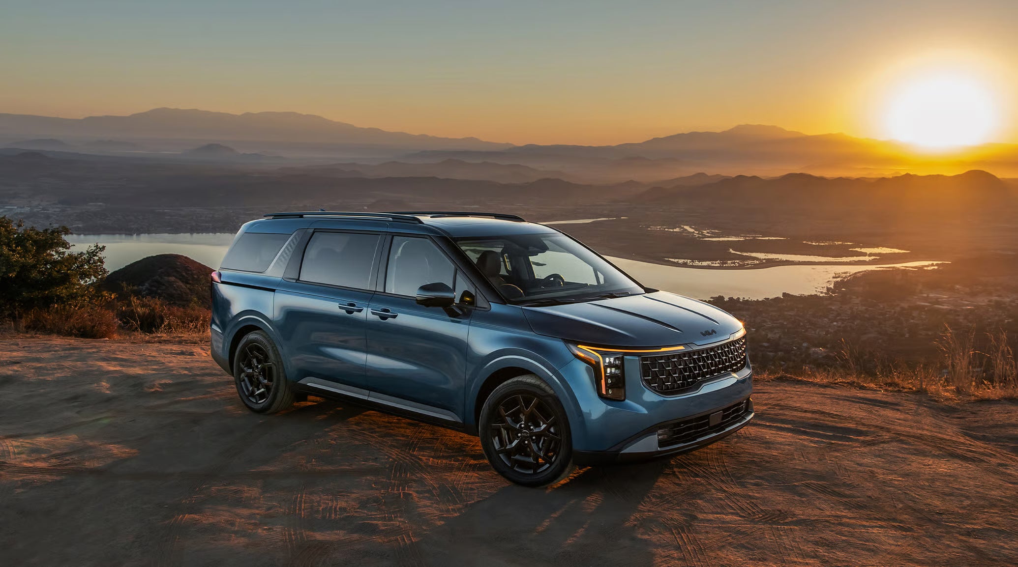A blue 2027 Kia Carnival parked on a mountain ledge at sunrise